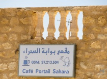 На фотографии изображена вывеска кафе 'Café Portail Sahara', расположенного на фоне стены из камня.