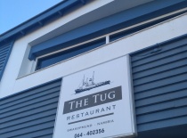 На фотографии видно вывеску ресторана 'The Tug', расположенного в Свакопмунде, Намибия. Здание выпол
