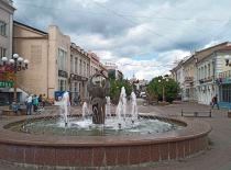 На фотографии изображена площадь с фонтаном и рядом зданий, вероятно, в центре города. Видны прохожи