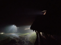 На фотографии изображен темный пейзаж с силуэтами хижин и лучами света, пробивающимися сквозь ночную