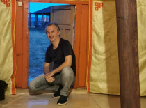 На фотографии изображен человек, сидящий на полу юрты с печью в центре. Видно, что юрта оформлена в
